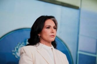 A head-and-shoulders portrait of Tulsi Gabbard, a woman with dark hair and a streak of gray, wearing a white blazer and looking to the side with a serious expression.