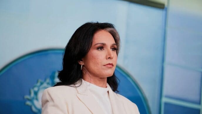 A head-and-shoulders portrait of Tulsi Gabbard, a woman with dark hair and a streak of gray, wearing a white blazer and looking to the side with a serious expression.