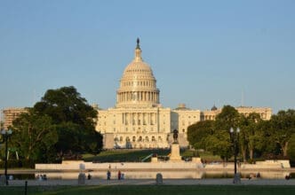 The U.S. Capitol building, with its grand dome and columns, stands majestically behind a reflecting pool and manicured lawn with trees and a statue, under a clear blue sky at sunset.