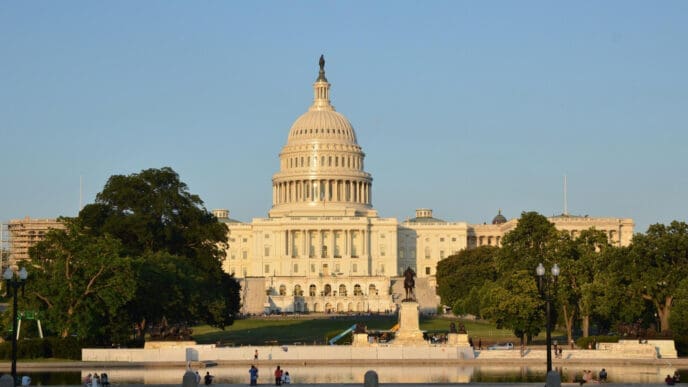 The U.S. Capitol building, with its grand dome and columns, stands majestically behind a reflecting pool and manicured lawn with trees and a statue, under a clear blue sky at sunset.