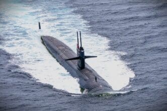 A wide, high-angle view of a large black US military submarine partially submerged, with its conning tower and periscopes visible, creating a white wake in the deep blue ocean.