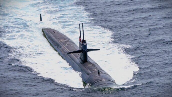 A wide, high-angle view of a large black US military submarine partially submerged, with its conning tower and periscopes visible, creating a white wake in the deep blue ocean.