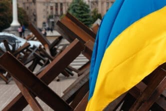 The Ukrainian flag hangs over metal barricades on a street in a Ukrainian city, with buildings and people in the blurry background.