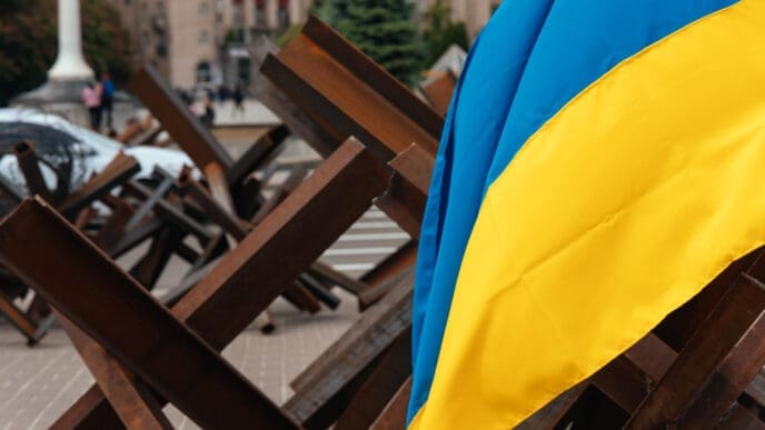 The Ukrainian flag hangs over metal barricades on a street in a Ukrainian city, with buildings and people in the blurry background.