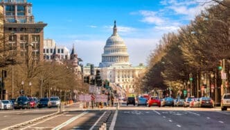 The United States Capitol building stands at the end of a busy street lined with trees and cars