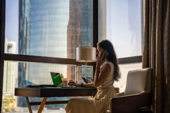A woman on a phone call, sitting at a desk with a laptop in a hotel room with a view of a city skyscraper.