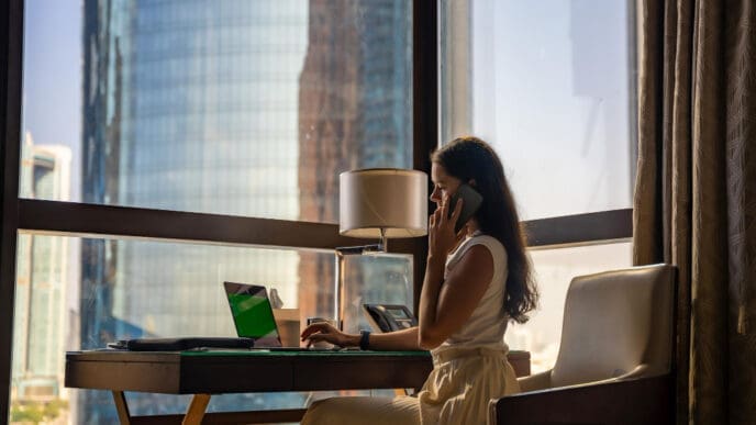A woman on a phone call, sitting at a desk with a laptop in a hotel room with a view of a city skyscraper.