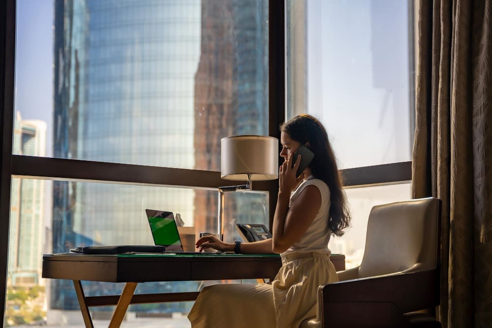 A woman on a phone call, sitting at a desk with a laptop in a hotel room with a view of a city skyscraper.