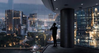 A silhouette of a millionaire on a smartphone stands by a large glass window, looking out at a stunning, illuminated city skyline and a river at dusk.