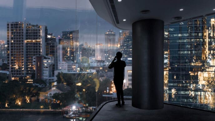A silhouette of a millionaire on a smartphone stands by a large glass window, looking out at a stunning, illuminated city skyline and a river at dusk.