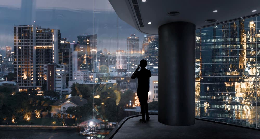 A silhouette of a millionaire on a smartphone stands by a large glass window, looking out at a stunning, illuminated city skyline and a river at dusk.