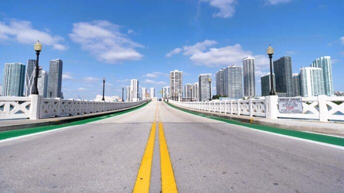 A low-angle shot looking down the Venetian Causeway road, with white decorative railings and a view of the Miami skyline on a sunny day.