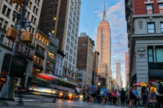 A long exposure photo of a bustling street in New York City, with the Empire State Building in the background and a bus and people blurred by motion.