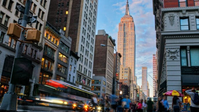 A long exposure photo of a bustling street in New York City, with the Empire State Building in the background and a bus and people blurred by motion.