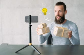 A young man with a beard records a video on his smartphone, holding up gift-wrapped boxes with an enthusiastic expression and a cartoon lightbulb above his head.