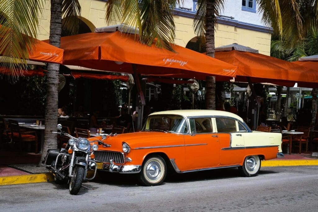 A vintage orange and cream-colored car is parked on a street next to a motorcycle, with a restaurant and palm trees visible behind it.