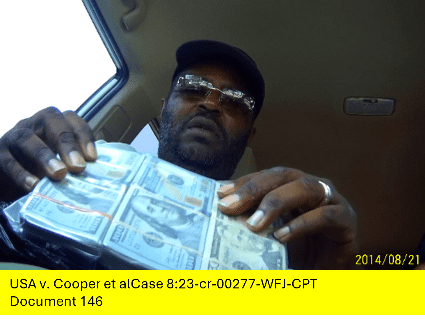 Virgil Cooper, sitting in a car, is shown in a photograph holding a large bundle of U.S. hundred-dollar bills.