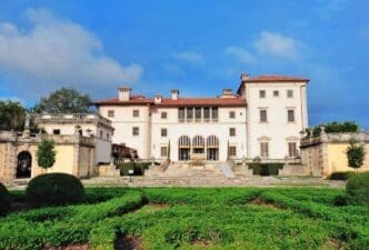 The historic Vizcaya Museum, a grand villa with red tile roofs and a formal garden, is seen under a blue sky with clouds.