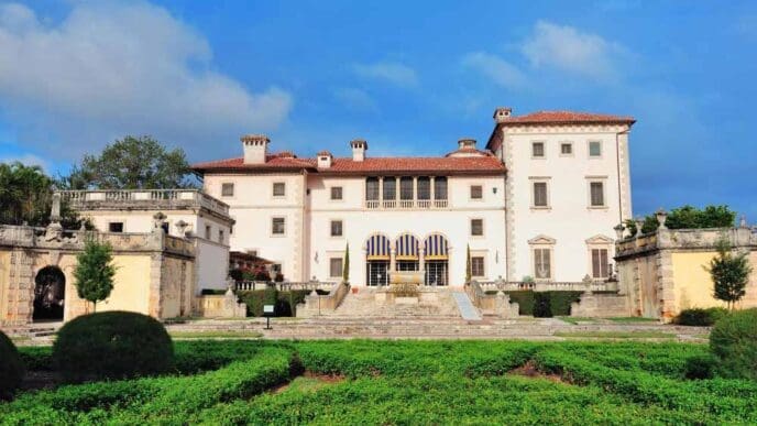 The historic Vizcaya Museum, a grand villa with red tile roofs and a formal garden, is seen under a blue sky with clouds.