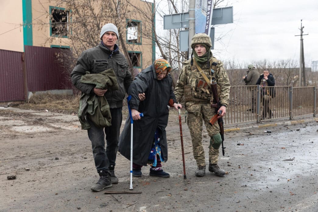 An elderly woman with a cane and a man holding a bag are standing on a muddy street next to a soldier in camouflage, with a damaged building in the background.