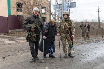 An elderly woman with a cane and a man holding a bag are standing on a muddy street next to a soldier in camouflage, with a damaged building in the background.