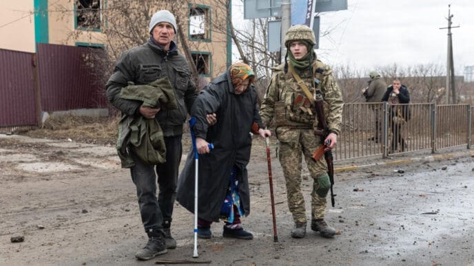 An elderly woman with a cane and a man holding a bag are standing on a muddy street next to a soldier in camouflage, with a damaged building in the background.