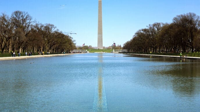 The Washington Monument is perfectly reflected in the Reflecting Pool, with a row of trees on each side under a clear blue sky.