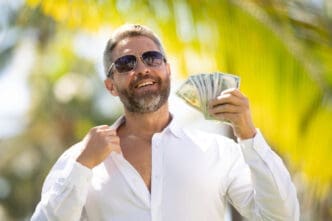 A smiling man with a beard and sunglasses holds a fan of US dollar bills while standing in front of blurry palm trees.