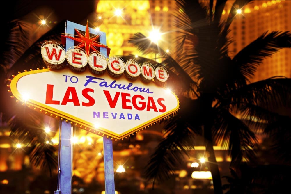 The iconic "Welcome to Fabulous Las Vegas Nevada" sign is brightly lit at night, with a palm tree and the city's lights blurred in the background.