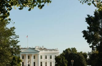 The White House is partially visible behind a frame of green trees against a clear blue sky