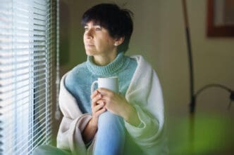 A serious woman with short dark hair, wrapped in a white blanket and a blue turtleneck sweater, sits and looks pensively out a window, holding a white mug.