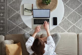 A woman, seen from a high angle, works on a laptop on a round white coffee table, sitting on a light-colored couch. A notebook, glasses, and a smartphone are also on the table.
