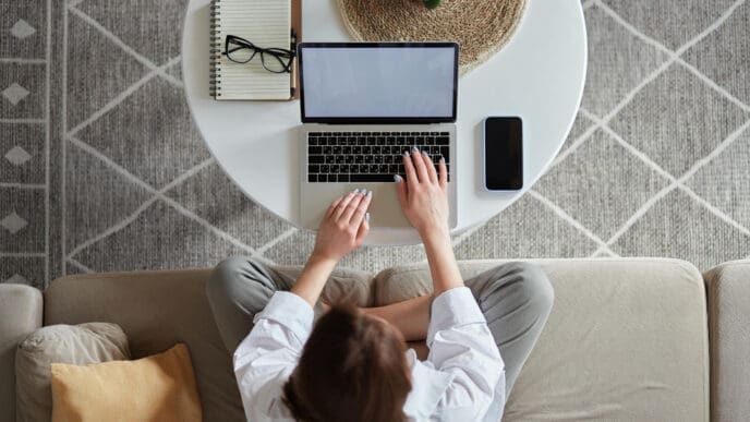 A woman, seen from a high angle, works on a laptop on a round white coffee table, sitting on a light-colored couch. A notebook, glasses, and a smartphone are also on the table.