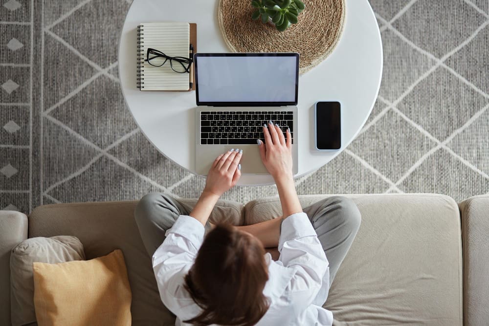 A woman, seen from a high angle, works on a laptop on a round white coffee table, sitting on a light-colored couch. A notebook, glasses, and a smartphone are also on the table.