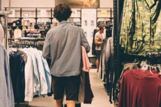 A young man, seen from behind, walks down a crowded aisle in a casual clothes store, carrying garments in his arms.