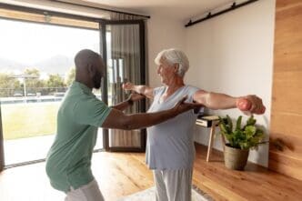 An African American male physiotherapist assists a Caucasian senior woman with dumbbell exercises.