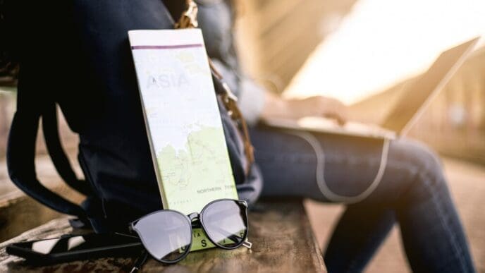 Asian woman wearing a hat and sunglasses uses a laptop while waiting at a train station, with a backpack, map, and earphones visible.