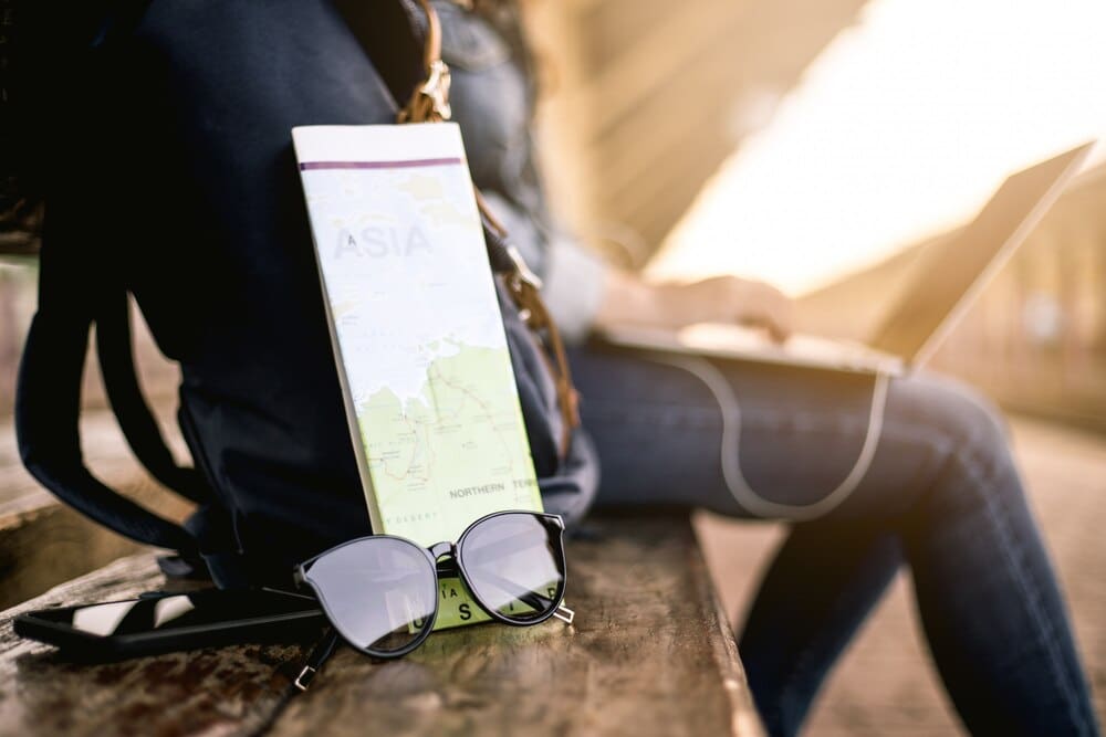 Asian woman wearing a hat and sunglasses uses a laptop while waiting at a train station, with a backpack, map, and earphones visible.
