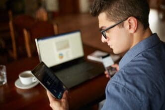 A man wearing glasses works with a laptop and smartphone, with charts displayed on the laptop screen, representing multitasking.