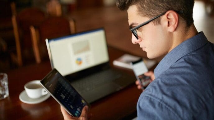 A man wearing glasses works with a laptop and smartphone, with charts displayed on the laptop screen, representing multitasking.