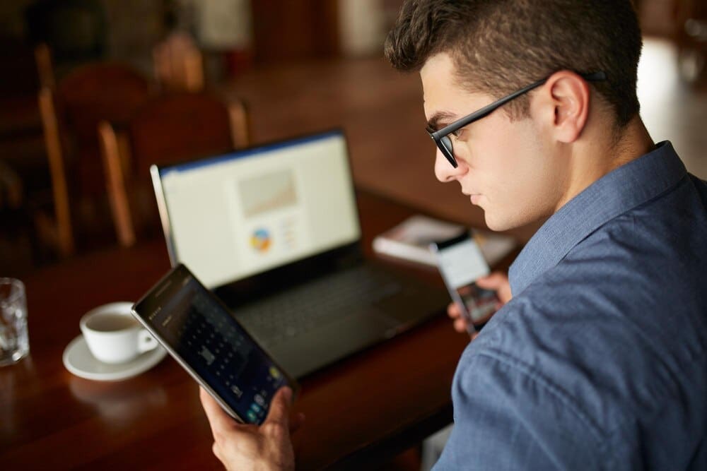 A man wearing glasses works with a laptop and smartphone, with charts displayed on the laptop screen, representing multitasking.