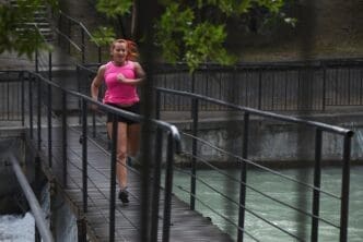 A red-haired woman in a pink top and shorts runs on a city bridge.