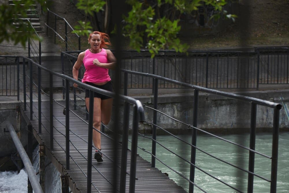 A red-haired woman in a pink top and shorts runs on a city bridge.