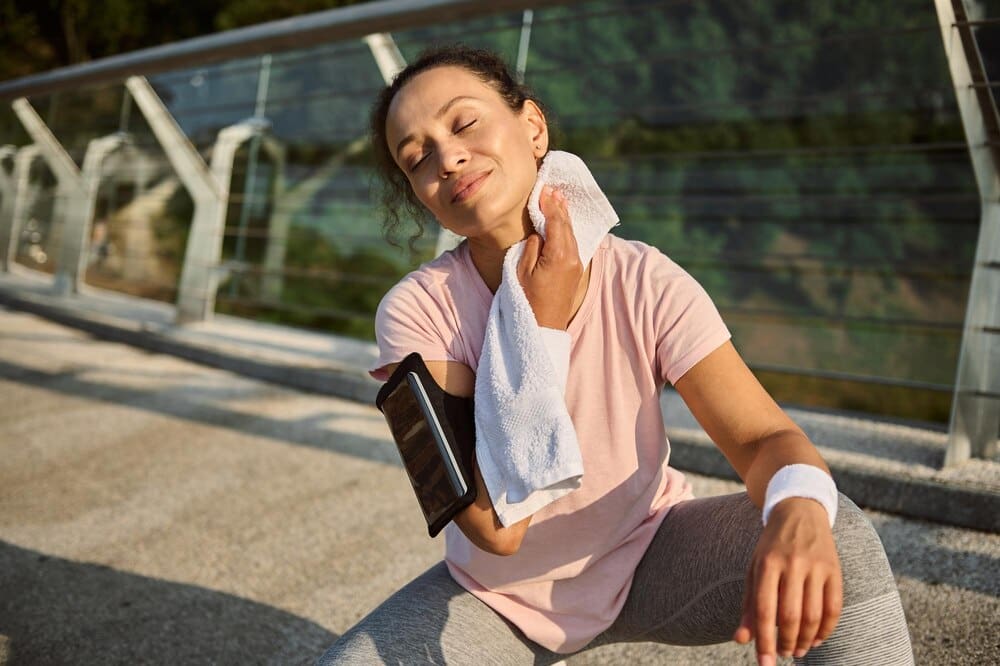 A smiling sportswoman wipes sweat from her face with a towel while resting on a city bridge after a morning jog.