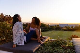 Biracial lesbian couple sits on a mattress in a garden at sunset, with copy space.