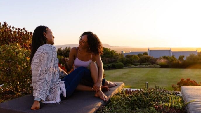 Biracial lesbian couple sits on a mattress in a garden at sunset, with copy space.