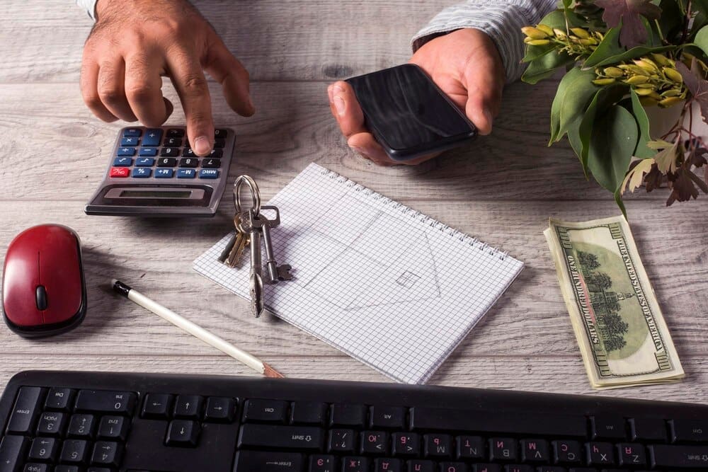 A businessman sits at a desk in an office, working on a laptop with documents spread across the table.