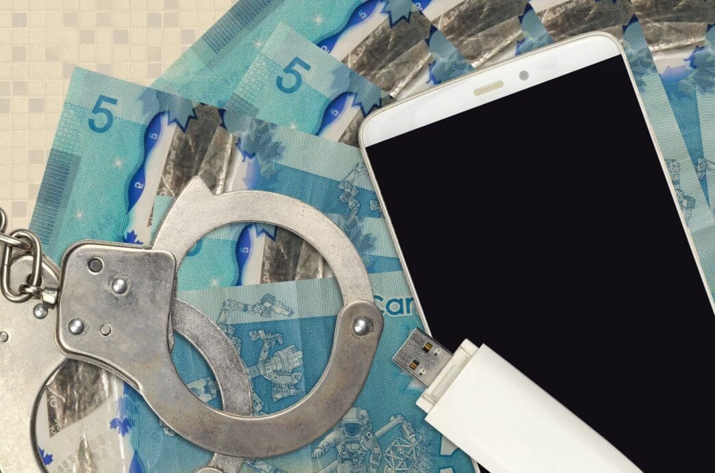 Canadian dollar bills and a smartphone are next to a pair of police handcuffs.