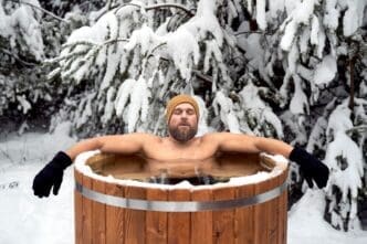A Caucasian man sits in a wooden tub outdoors in a snowy forest.