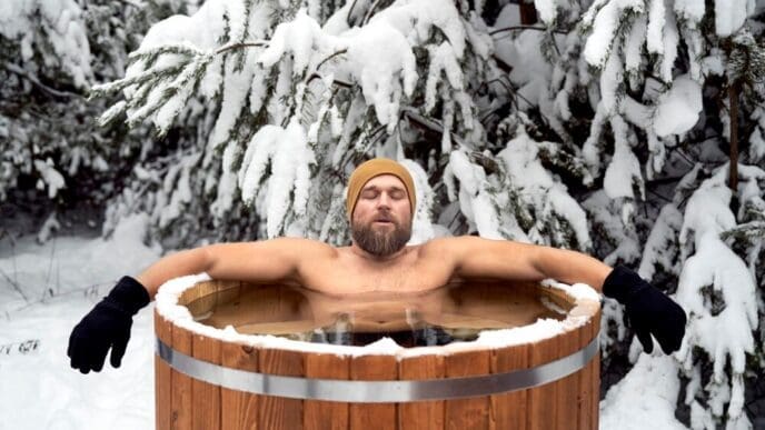 A Caucasian man sits in a wooden tub outdoors in a snowy forest.